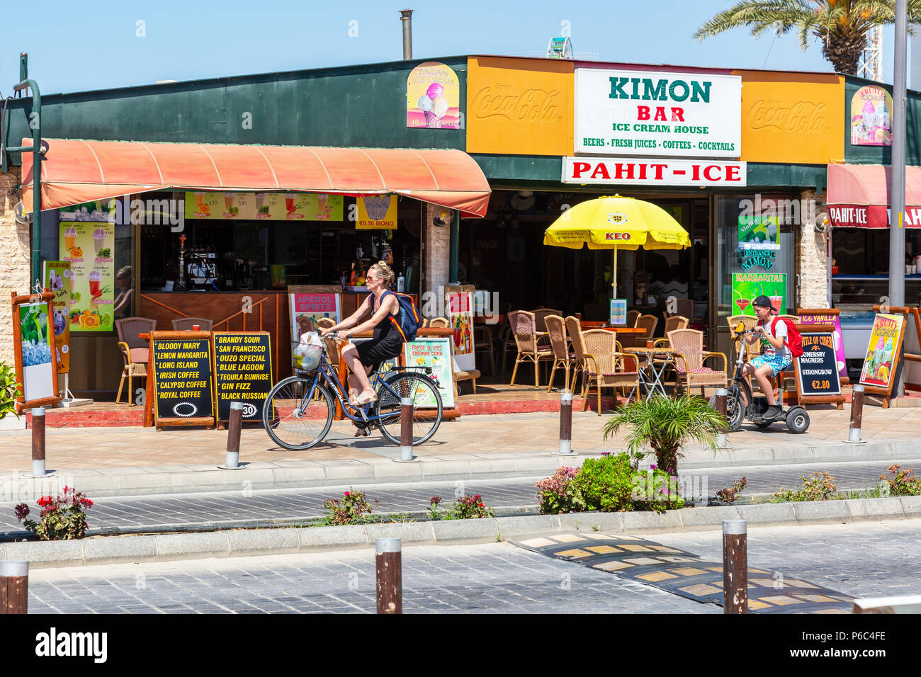 Woman on a bicycle followed by her son on an electric trike, cycling on a cycle track in front of a public bar, Ayia Napa, Cyprus Stock Photo