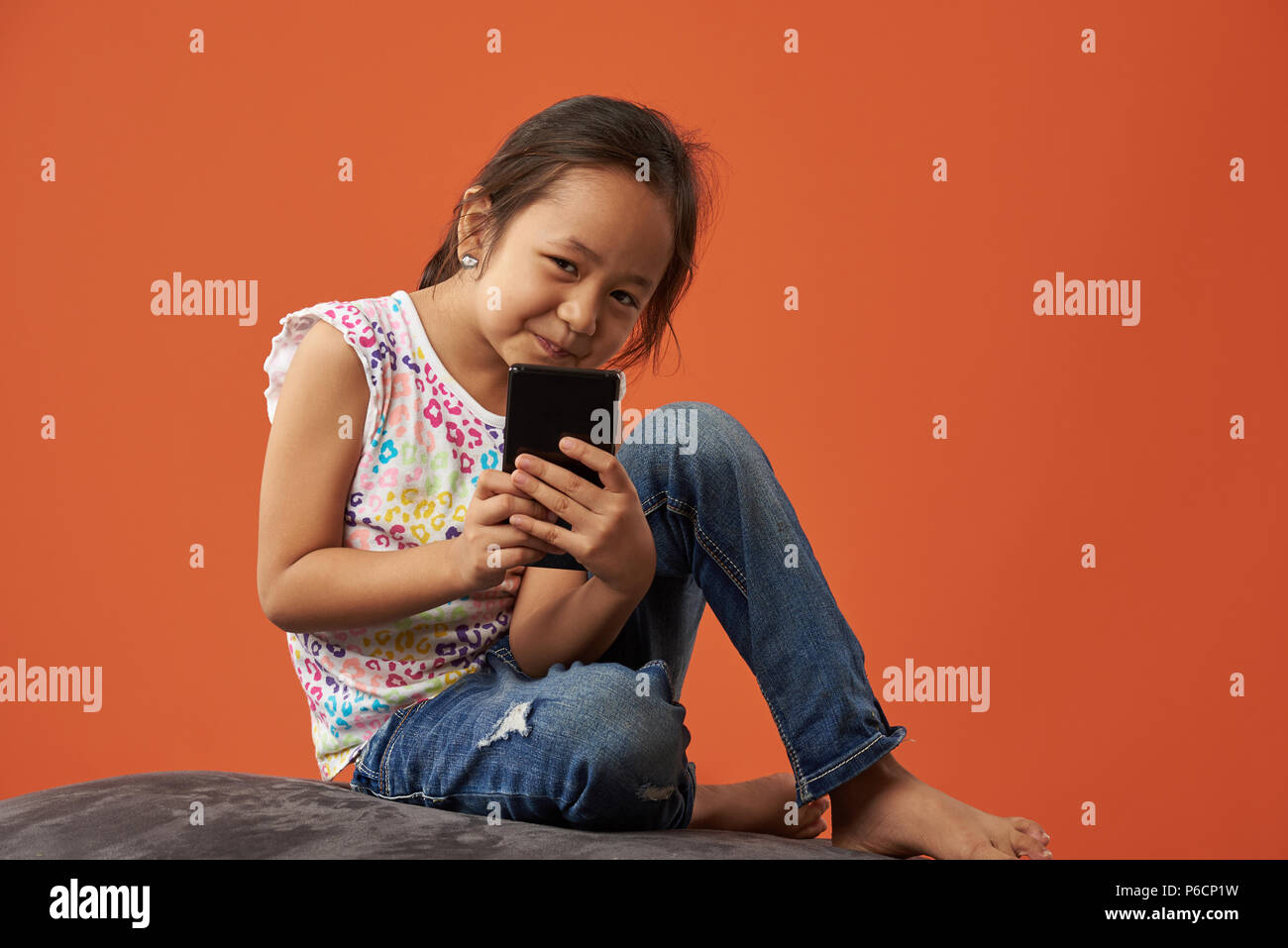 Asian little girl hold smartphone sit on sofa isolated on orange background Stock Photo