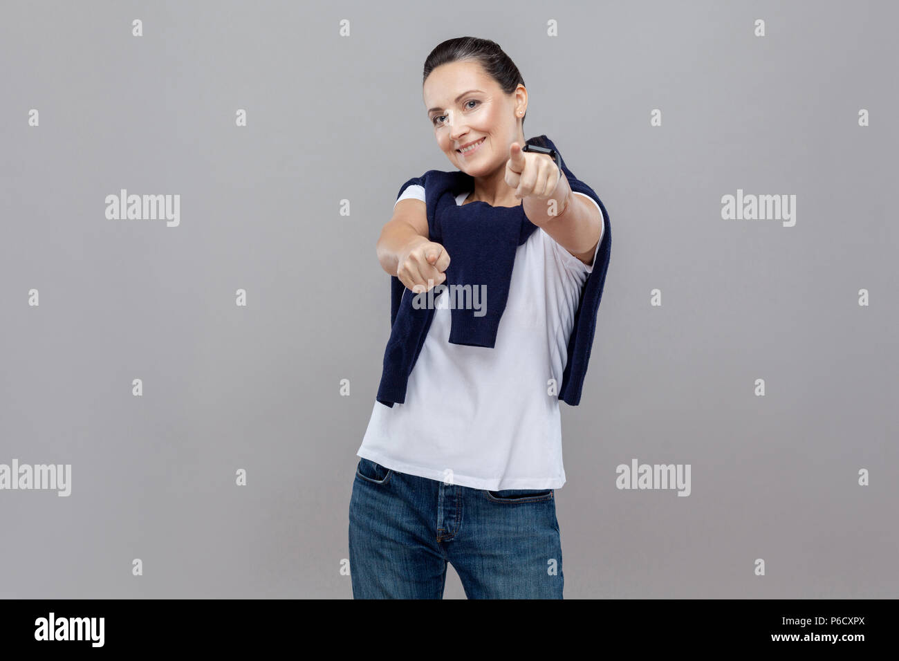 Adult cute woman in casual clothes choice fingers at camera with jeans and sweater on her shoulders, smart watch on arm, Studio shot, isolated on grey Stock Photo