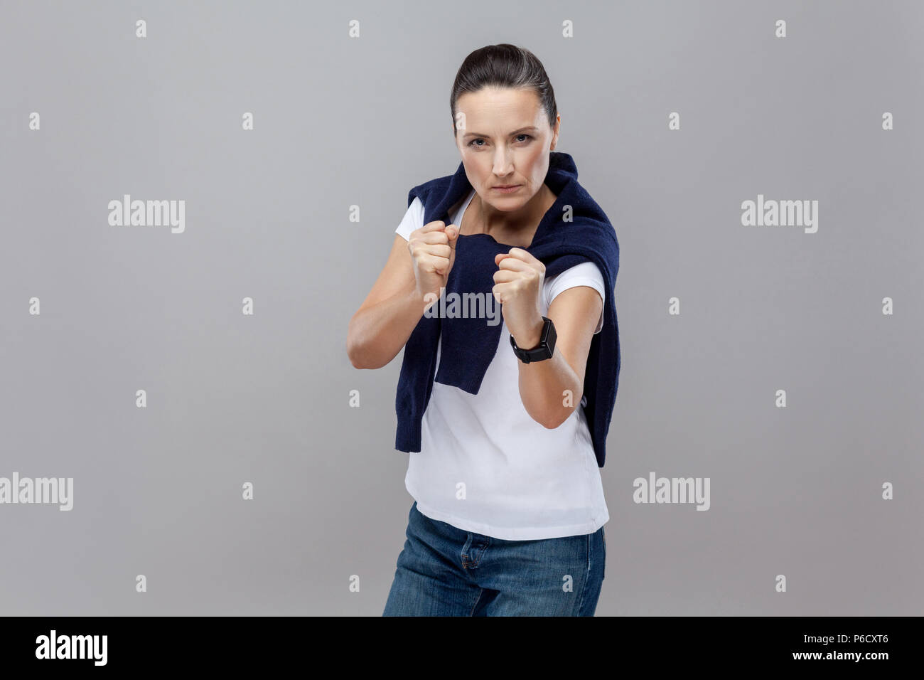 Boxing. Adult woman in casual clothes with blue jeans and sweater on her shoulders, smart watch on arm, ready for fight on grey background. isolated.  Stock Photo