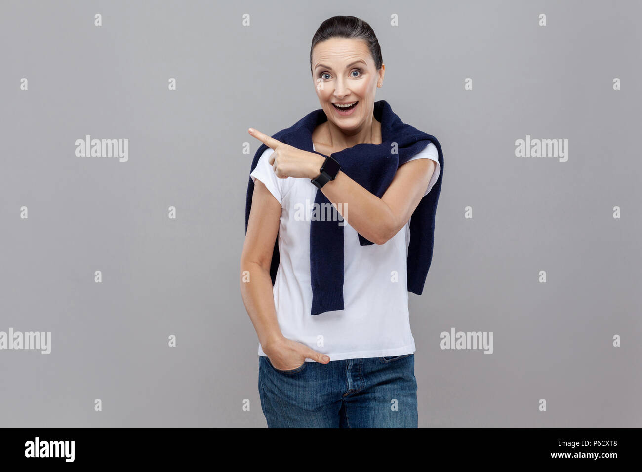 Look this! Hand finger pointing isolated on grey background with blue jeans and sweater on her shoulders, smart watch on arm,. Isolated studio shot Stock Photo