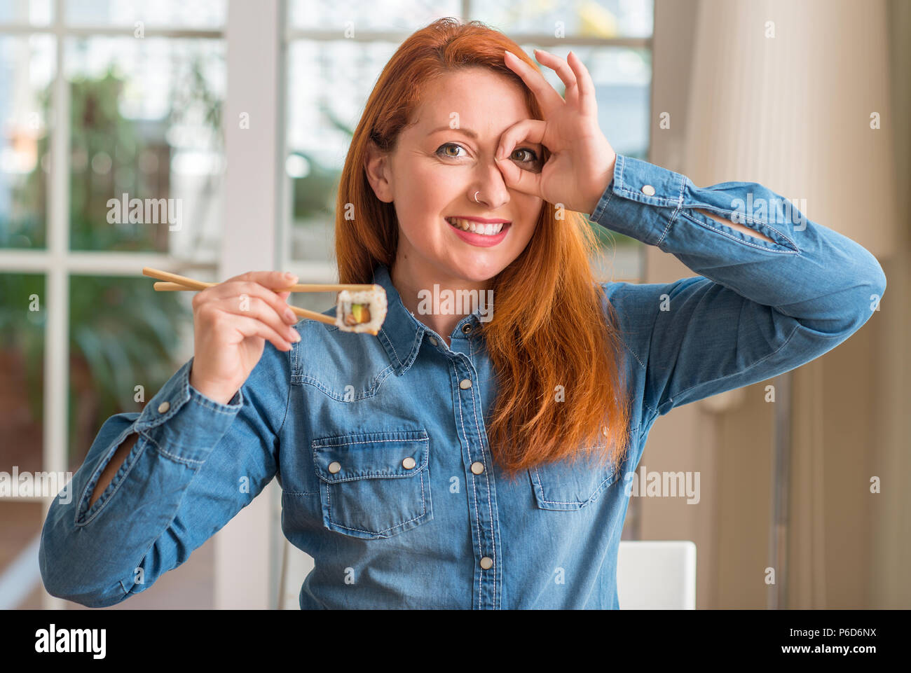 Redhead woman eating sushi using chopsticks with happy face smiling doing ok sign with hand on