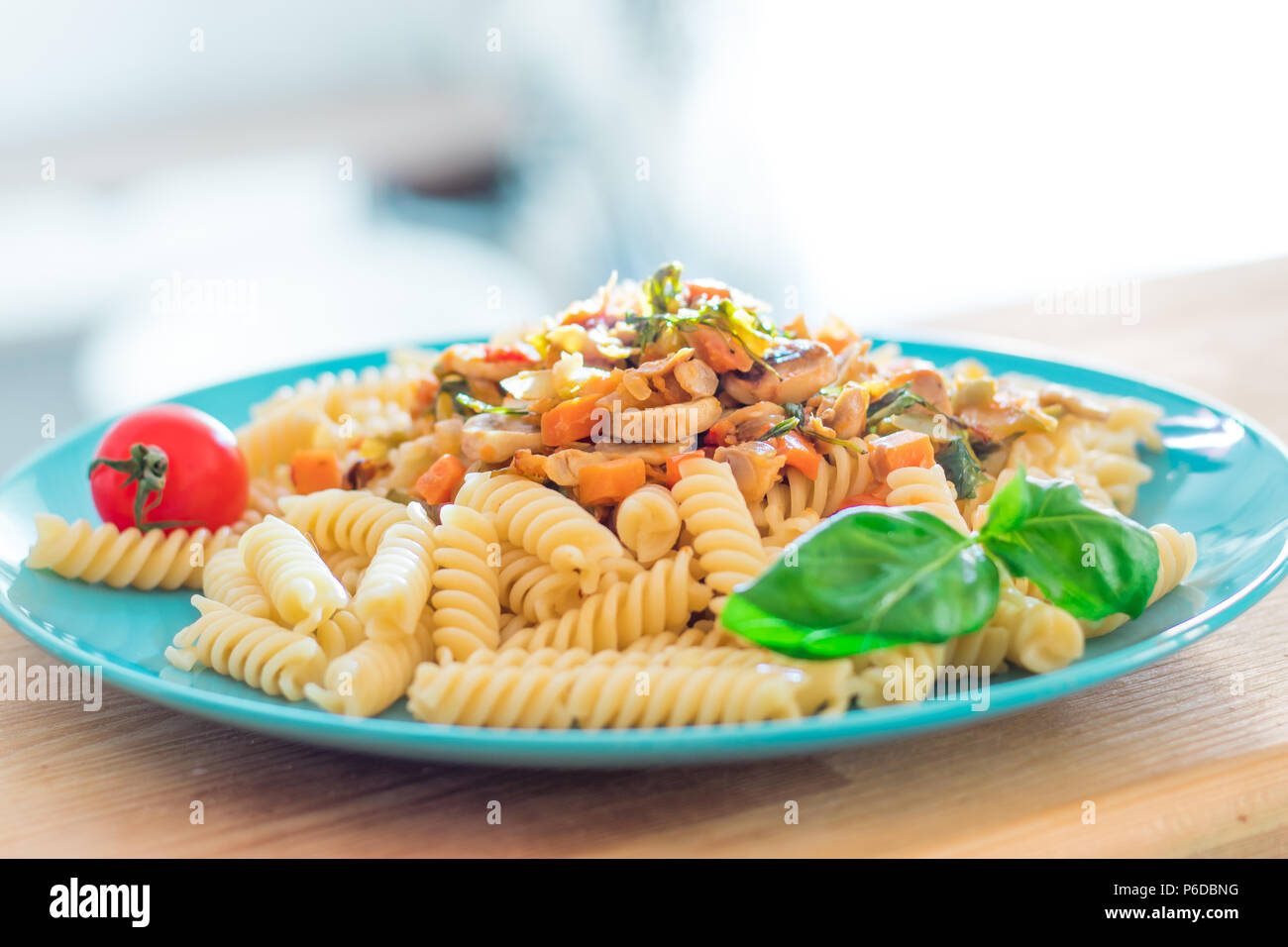 Cooked italian pasta fusilli with fried mushrooms, vegetables and fresh tomato cherry on turquoise plate and wooden table Stock Photo