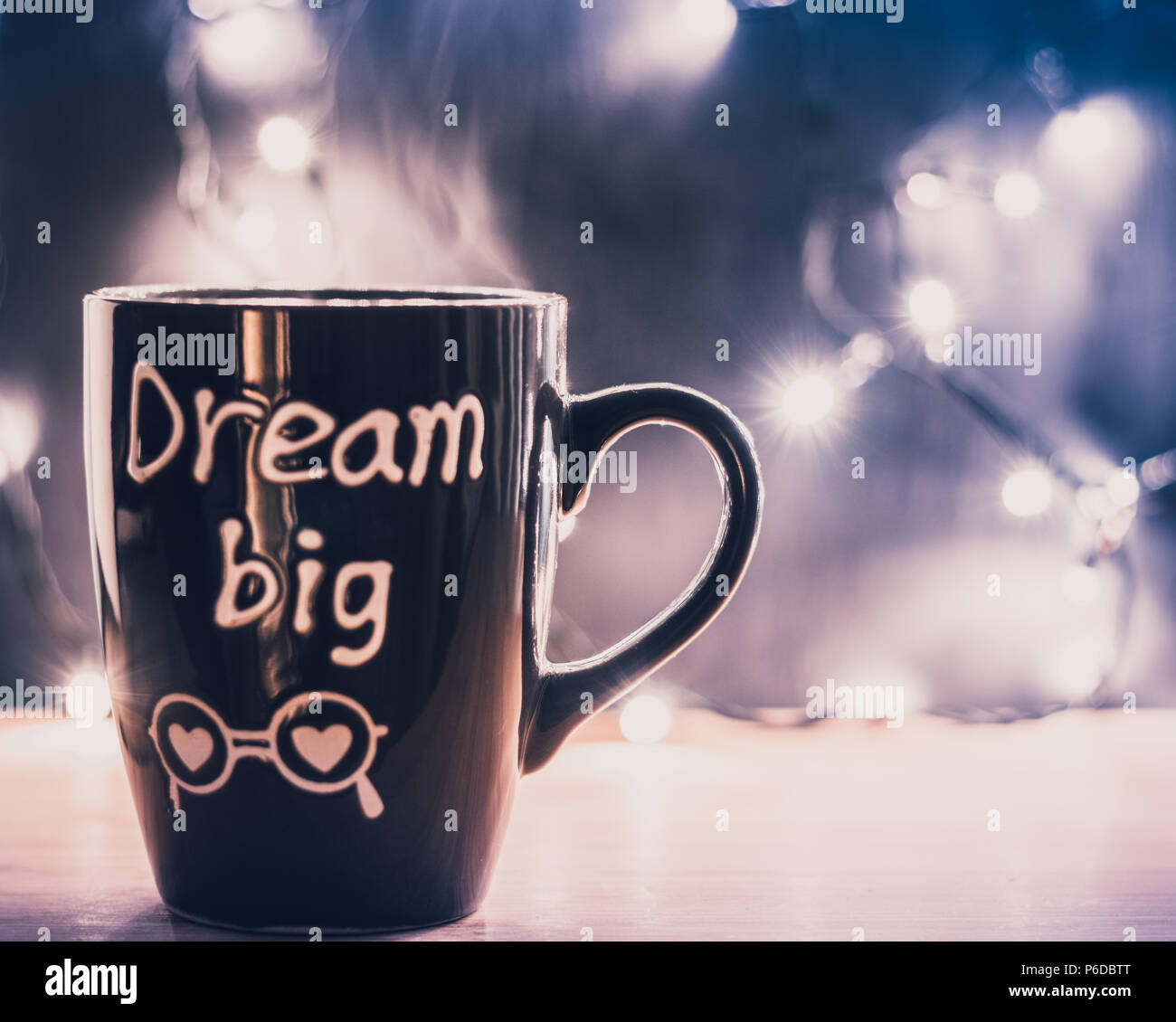 Coffee mug with letters dream big with steam on a wooden top and christmas small lights on a dark background. Copyspace. Front view Stock Photo