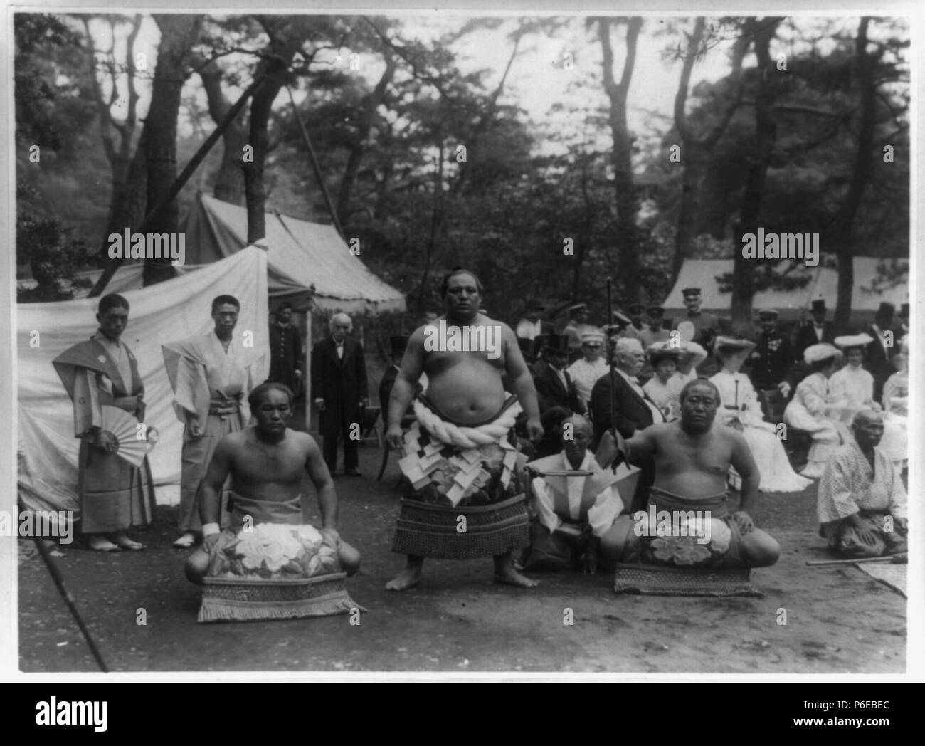 English: Three sumo wrestlers posed outdoors with spectators in background, probably in Japan, in approximately 1905. The middle wrestler Hitachiyama wears the distinctive belt indicating yokuzuna rank. circa 1905 75 Sumo wresters (1905) Stock Photo
