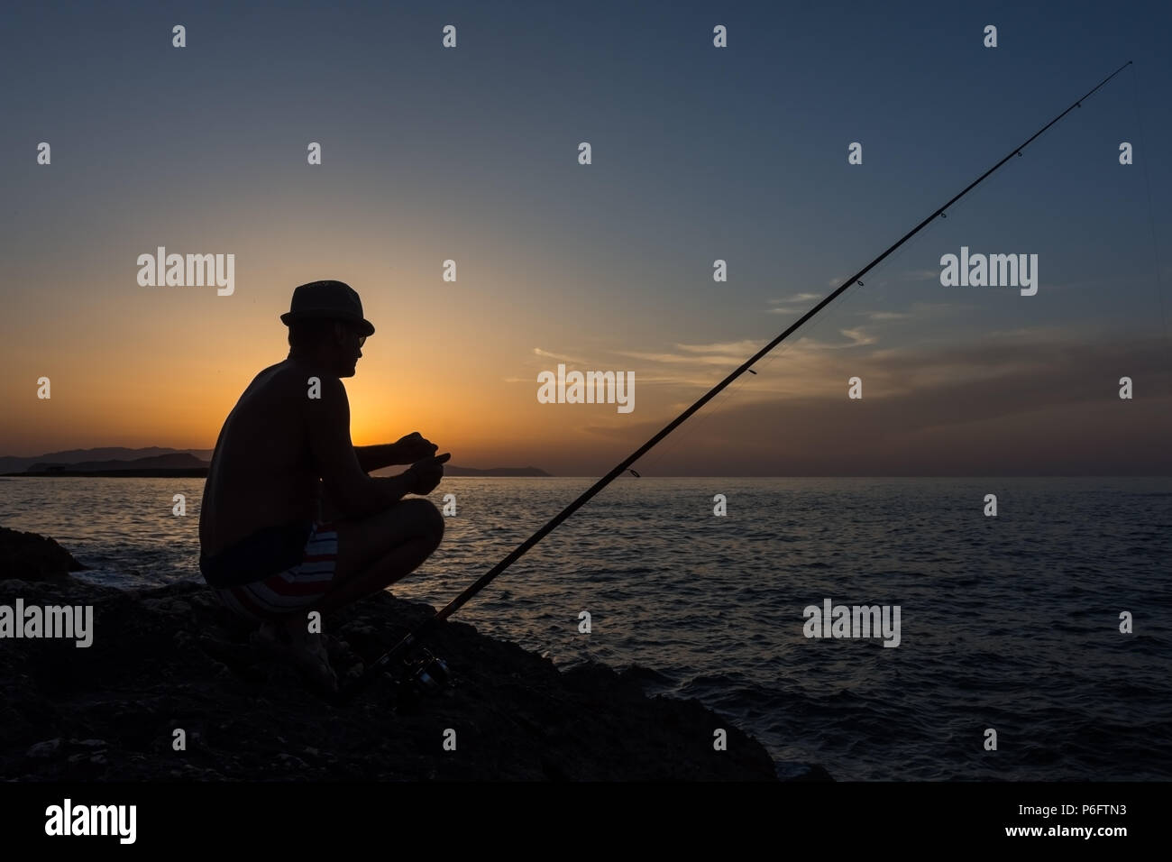 An adult man fishing on the rocks at sunset on the island of Crete in Greece Stock Photo