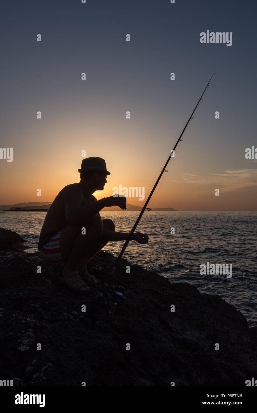 An adult man fishing on the rocks at sunset, on the seashore in Greece Stock Photo