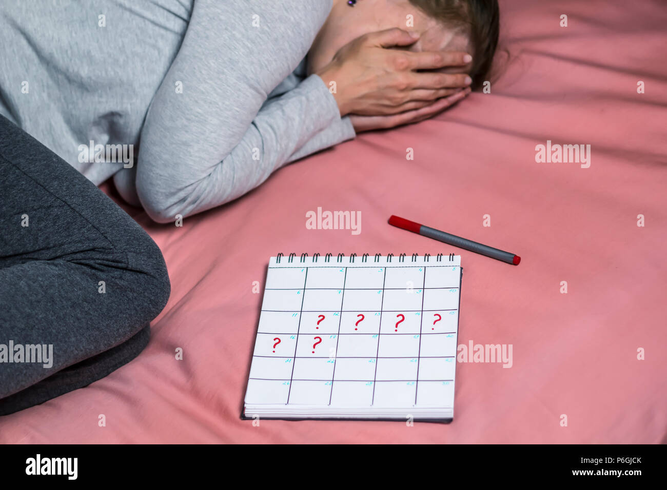 The calendar period with marked question marks lies near the young sad woman. A depressed girl, an unplanned pregnancy. Delay of menstruation. Stock Photo