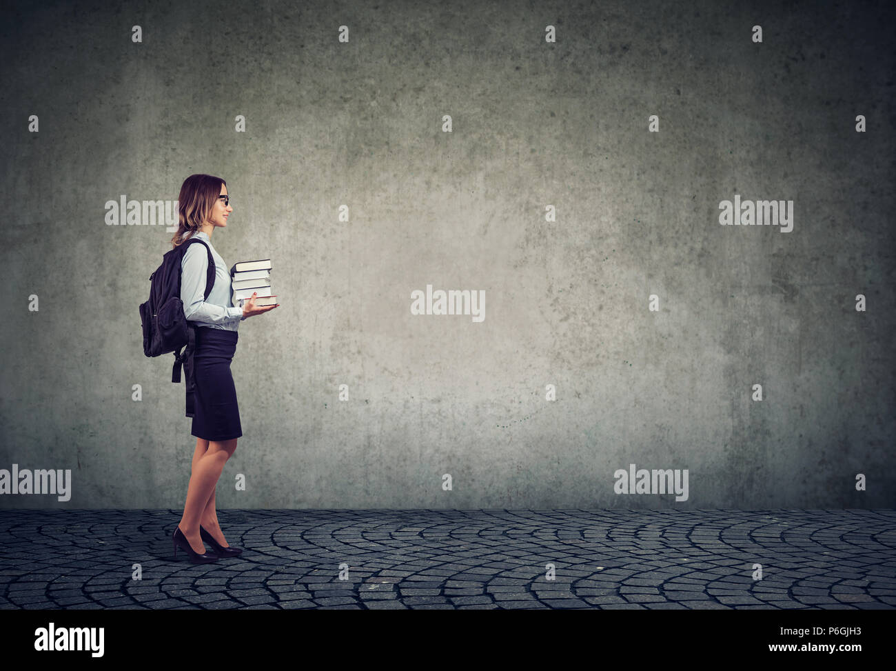 Side view of woman having studies in university and standing with stack of books and backpack against gray background Stock Photo