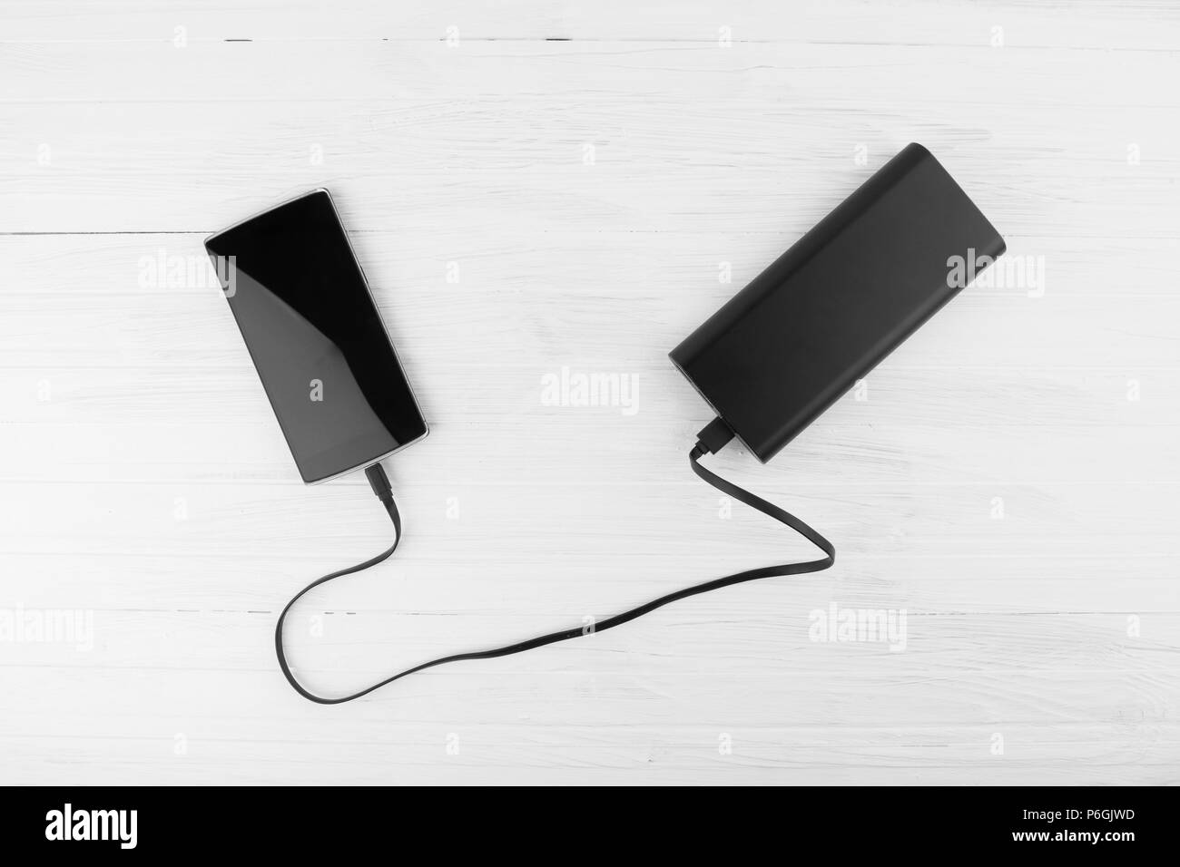 Black Smart Phone Connected To Big Battery Power Bank Charging Concept On White Painted Wooden Background Stock Photo