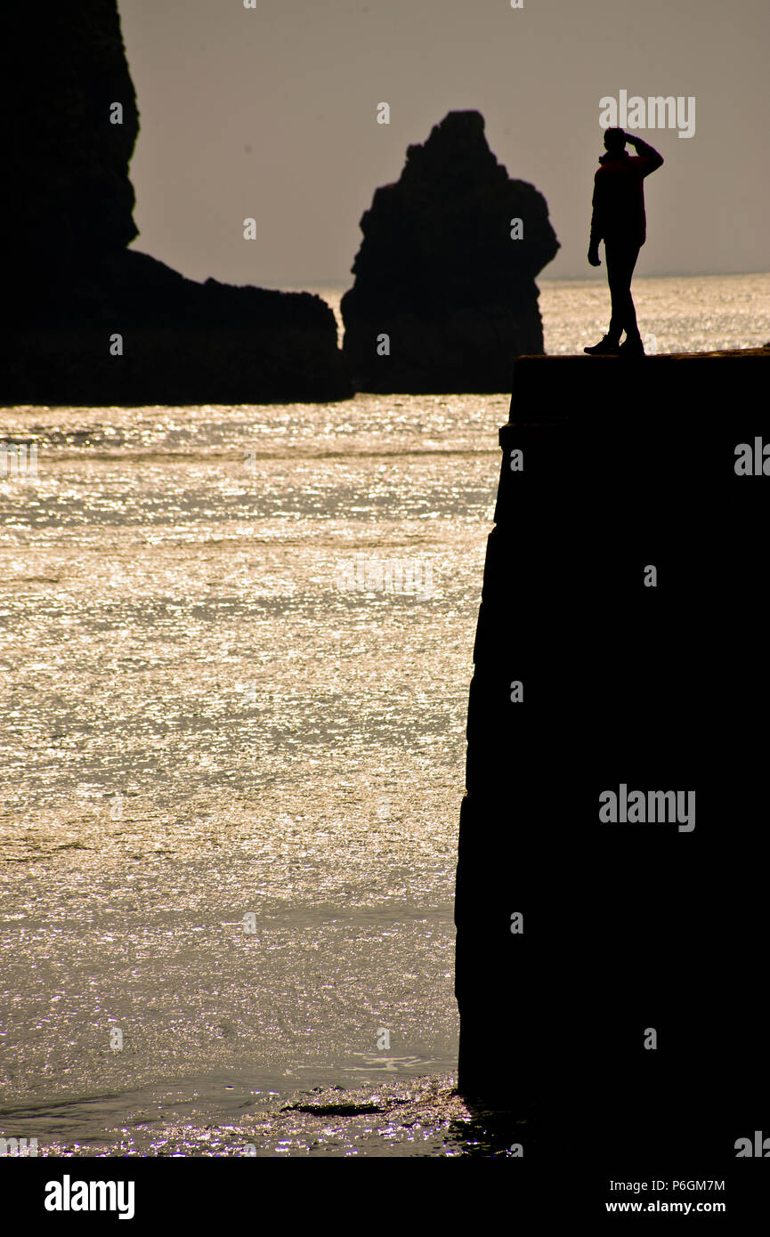A man standing on a Cornish seawall at sunset looking out to sea silhouetted against the sky Stock Photo
