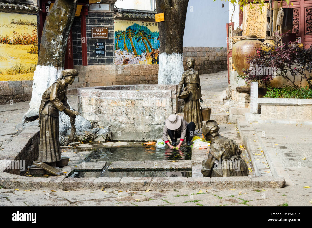 Local resident washing vegetables from a well next to three bronze statues of Naxi women in Lijiang Old Town, China Stock Photo