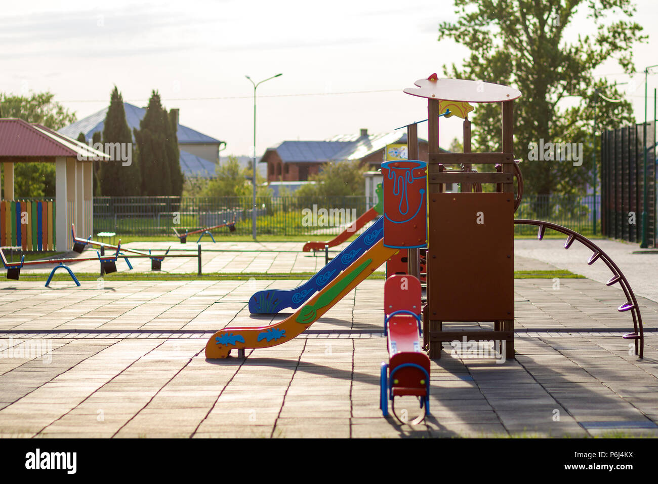 Beautiful playground in kindergarten with bright new alcove with red tile roof and multicolored low fence, green lawn, swings, slides and sandbox. Per Stock Photo