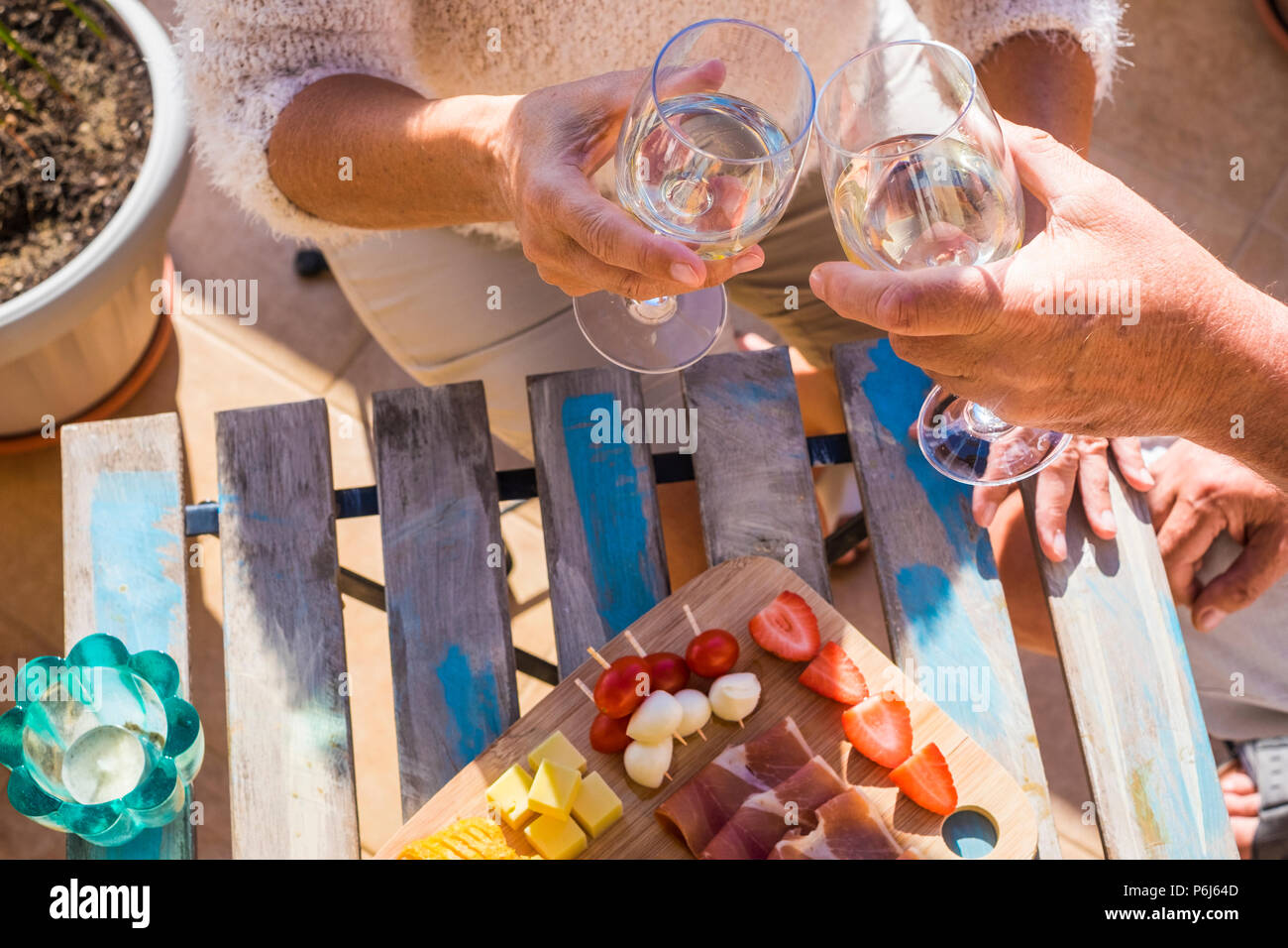 close up of two hands, a pair, from adult aged caucasian people do cheers with white wine and eat some fruits and snacks on a wood table outdoor under Stock Photo