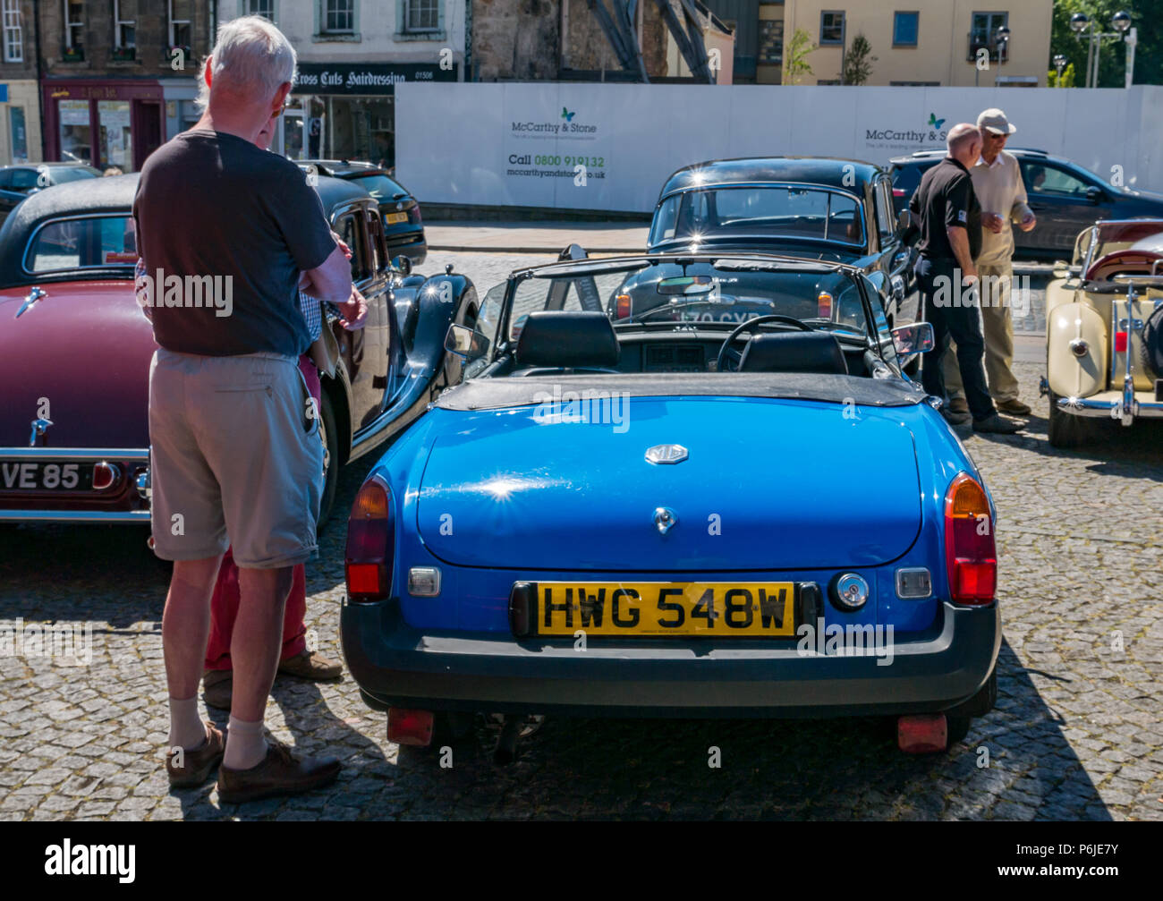 The Cross, Kirkgate, Linlithgow, West Lothian, Scotland, United Kingdom, 30th June 2018. Enthusiasts display sparkling vintage cars at a rally  on a sunny Summer day. People admiring a 1980 blue MG sports car Stock Photo