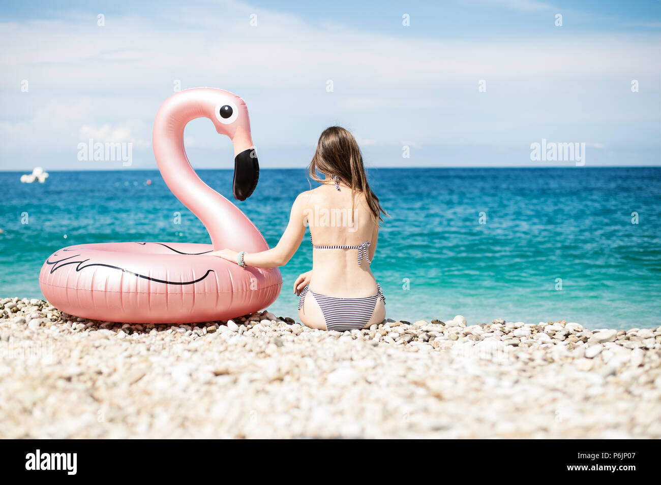 Young woman with bikini sitting close to giant inflated flamingo on a beach with turquois water of Ionian Sea Albania Stock Photo