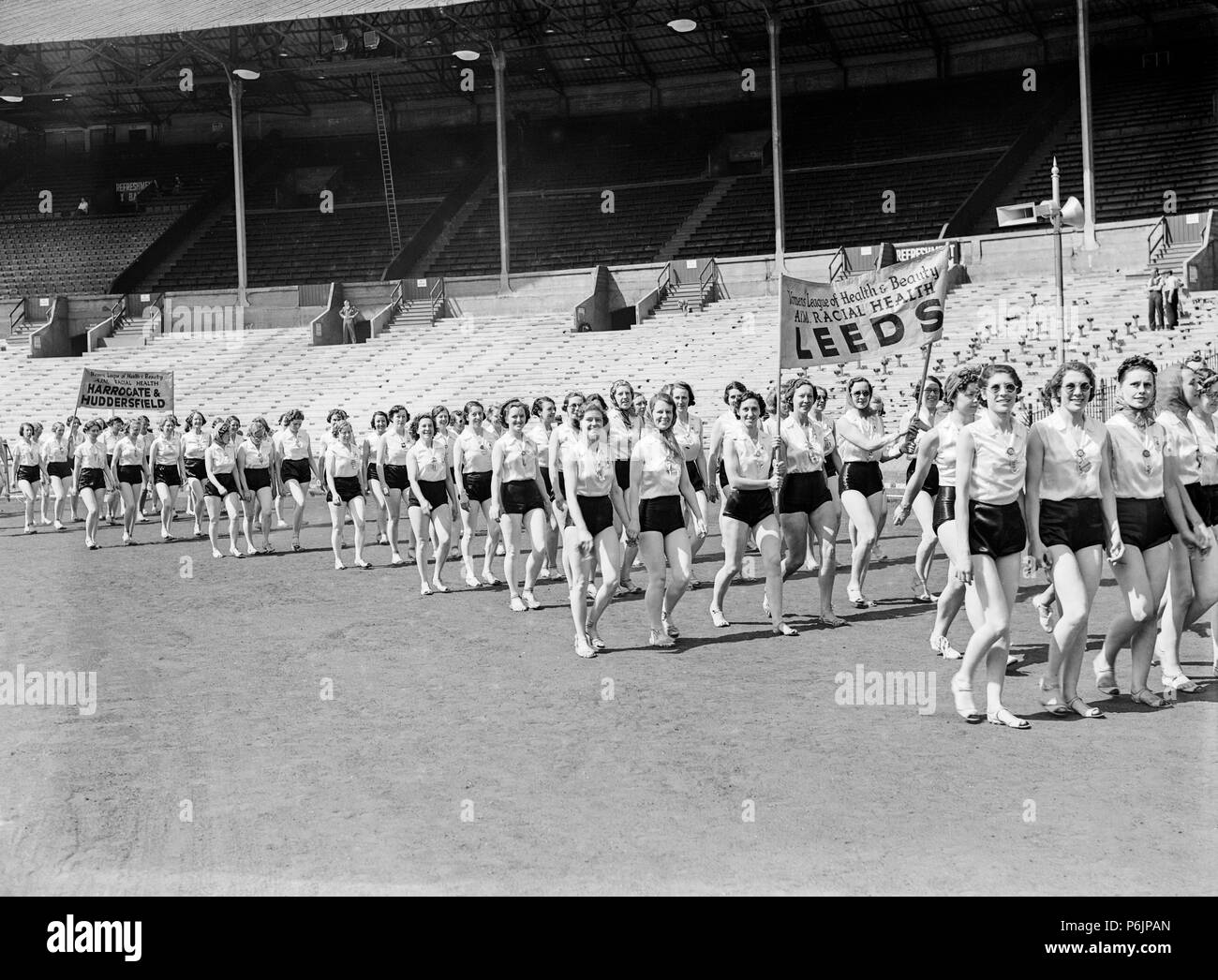 Members of The Women's League of Health and Beauty, marching inside a large sports stadium in England during the 1930s. Banners from the branches at Leeds, and Harrogate and Huddersfield are visible. They are campaigning about Racial Health. Stock Photo