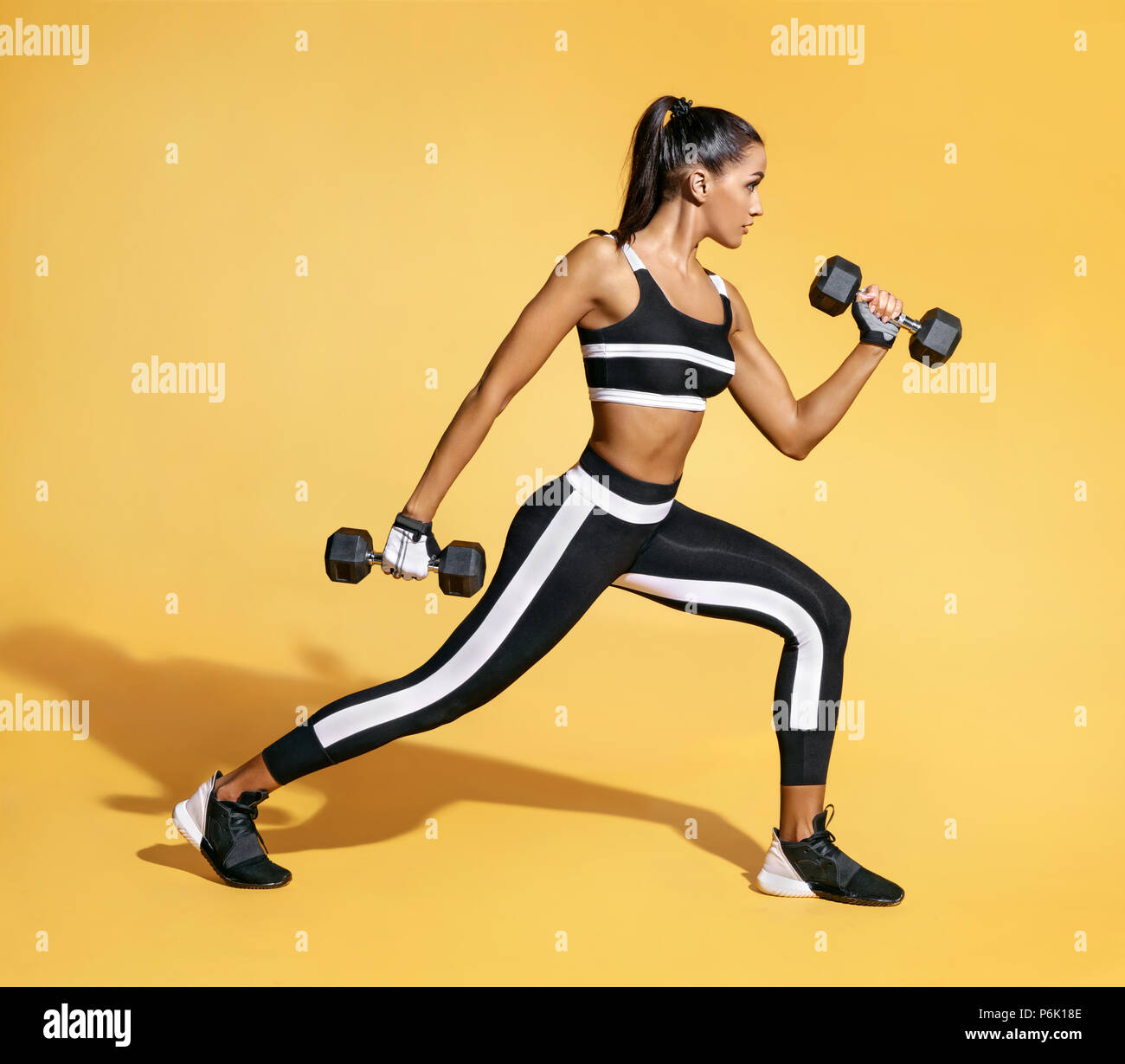 Sporty latin woman training muscles of hands and legs using a dumbbells. Photo of muscular woman in black sportswear on yellow background. Strength an Stock Photo