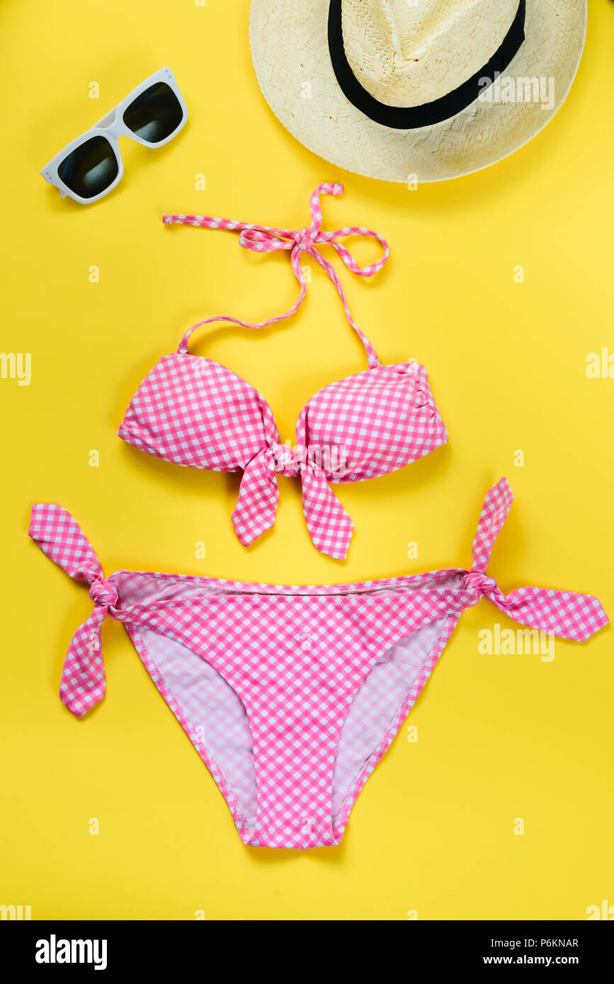 Top view of two pieces pink chequered swimming suit, white sunglasses and straw hat over yellow background. Stock Photo
