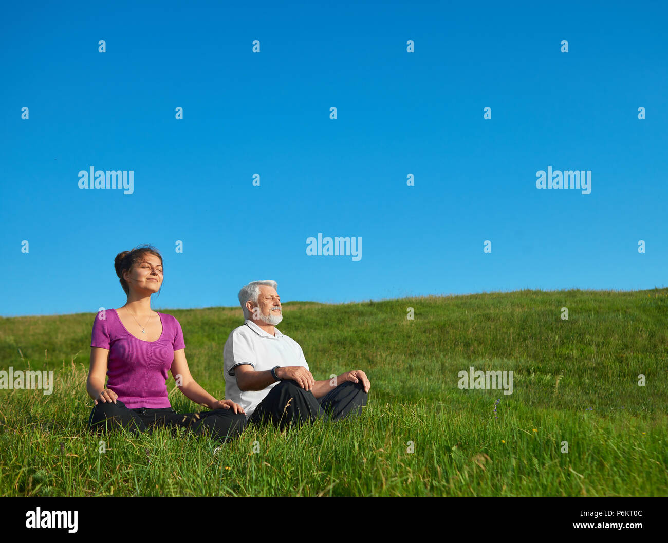 Young girl and old man meditating sitting in the field. Sitting in lotus pose on bright saturated sky background. Looking positive, satisfied, feeling good, concentrated. Calm and silent. Stock Photo