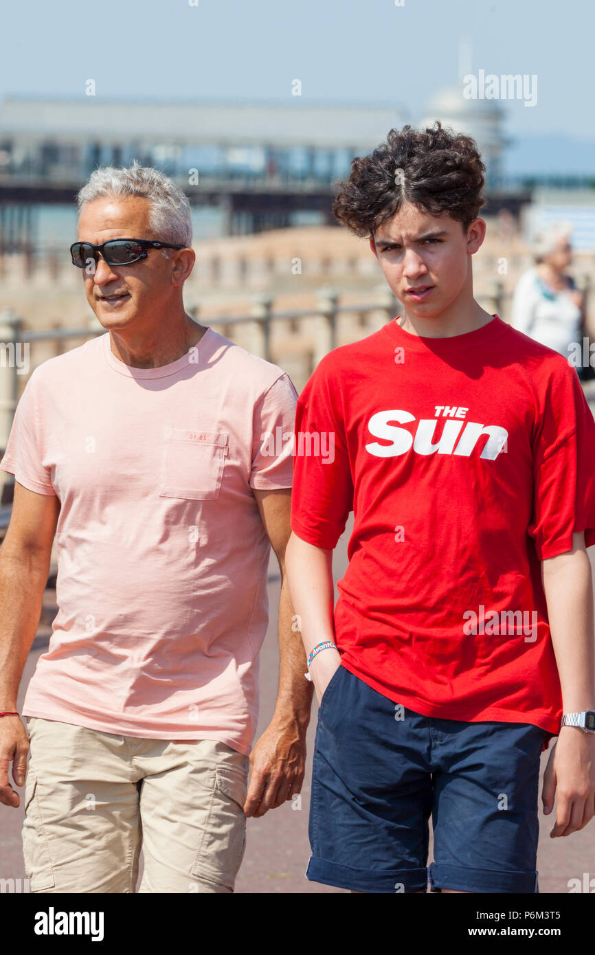 Hastings, East Sussex, UK. 1st Jul, 2018. UK Weather: The heatwave continues in Hastings with temperatures expected to exceed 24°C. Plenty of people are out and about walking along the seafront as the tide is out at the moment. This father and son visitors to Hastings with the young man wearing a 'The Sun' newspaper t shirt. Photo Credit: Paul Lawrenson / Alamy Live News Stock Photo