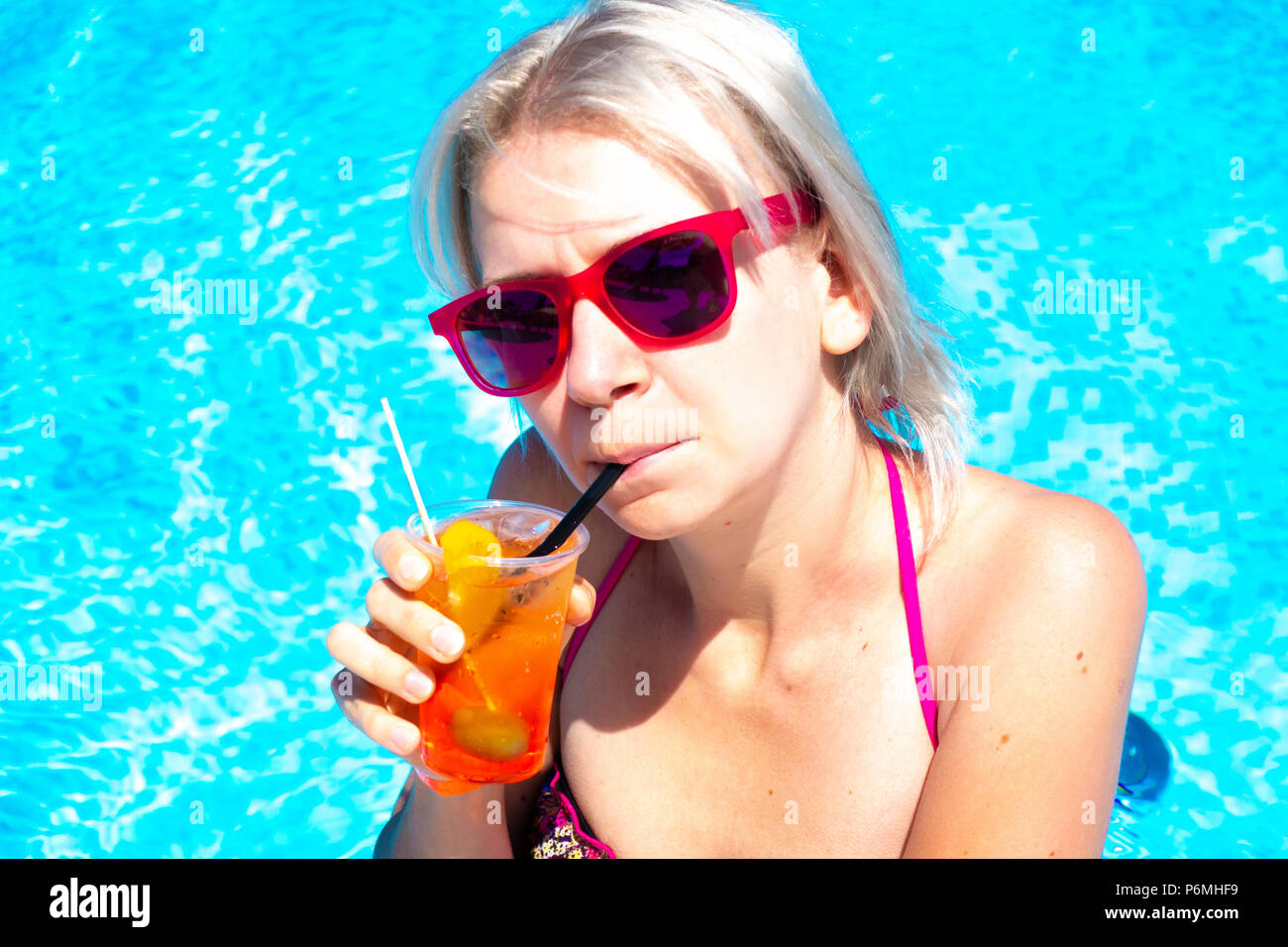 Young blonde woman with cocktail drink in pool. Happy gorgeous female with stylish sunglasses and bikini drinking cocktail and relaxing in the swimmin Stock Photo