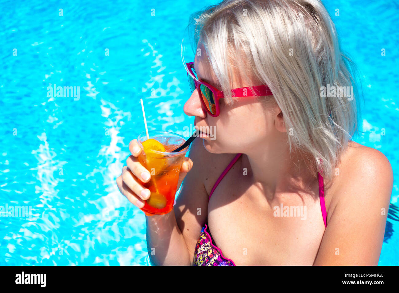 Young blonde woman with cocktail drink in pool. Happy gorgeous female with stylish sunglasses and bikini drinking cocktail and relaxing in the swimmin Stock Photo