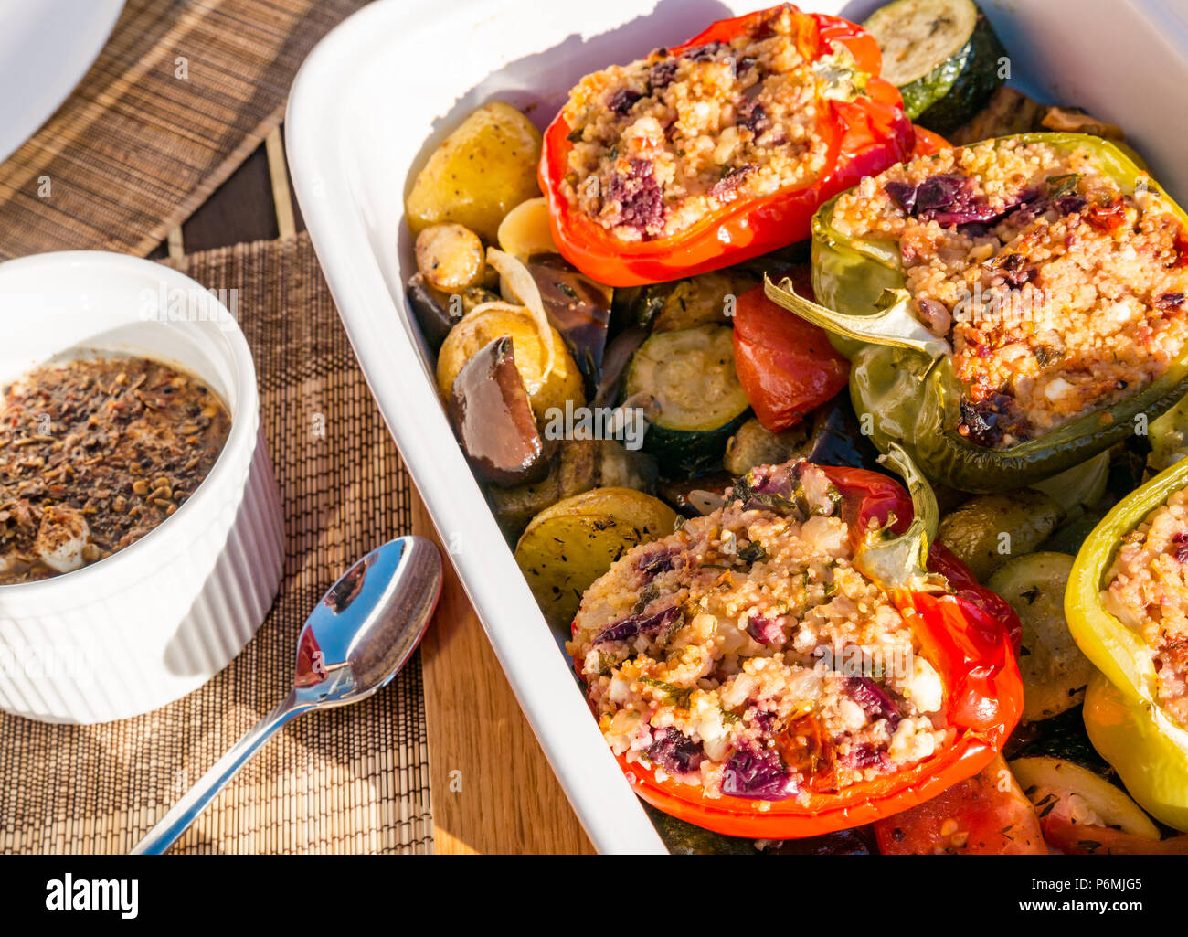 Stuffed roasted peppers in ceramic dish on a bed of roasted vegetables and potatoes with a spiced yoghurt dressing on outdoor table in sunshine Stock Photo