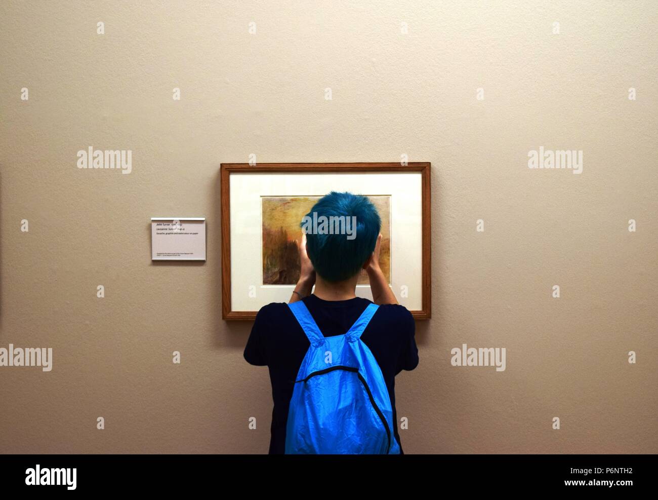 Young person with blue hair and matching rucksack takes close-up picture of Turner painting at Tate Britain. Stock Photo