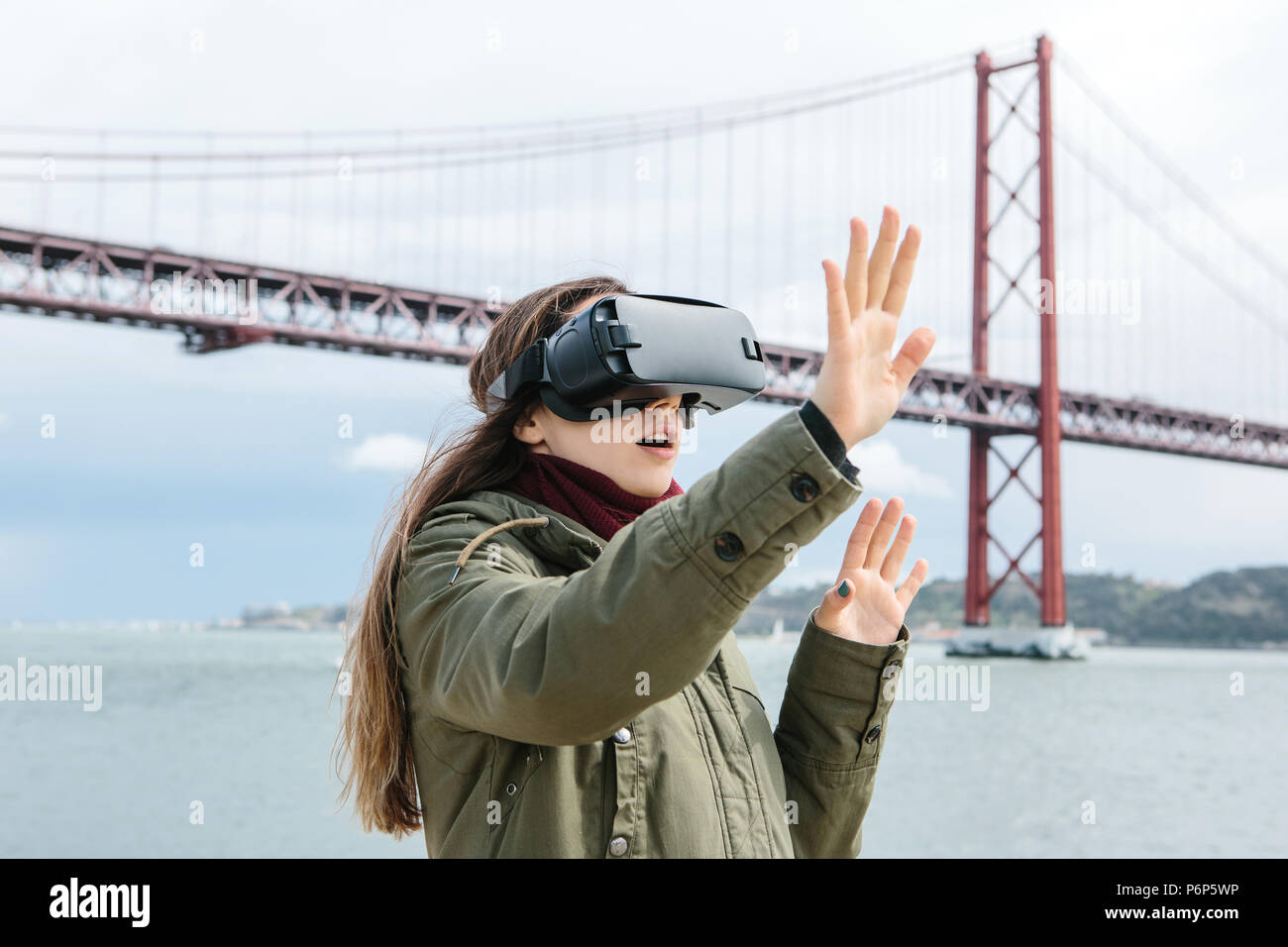 Young beautiful girl wearing virtual reality glasses. 25th of April bridge in Lisbon in the background. The concept of modern technologies and their use in everyday life Stock Photo