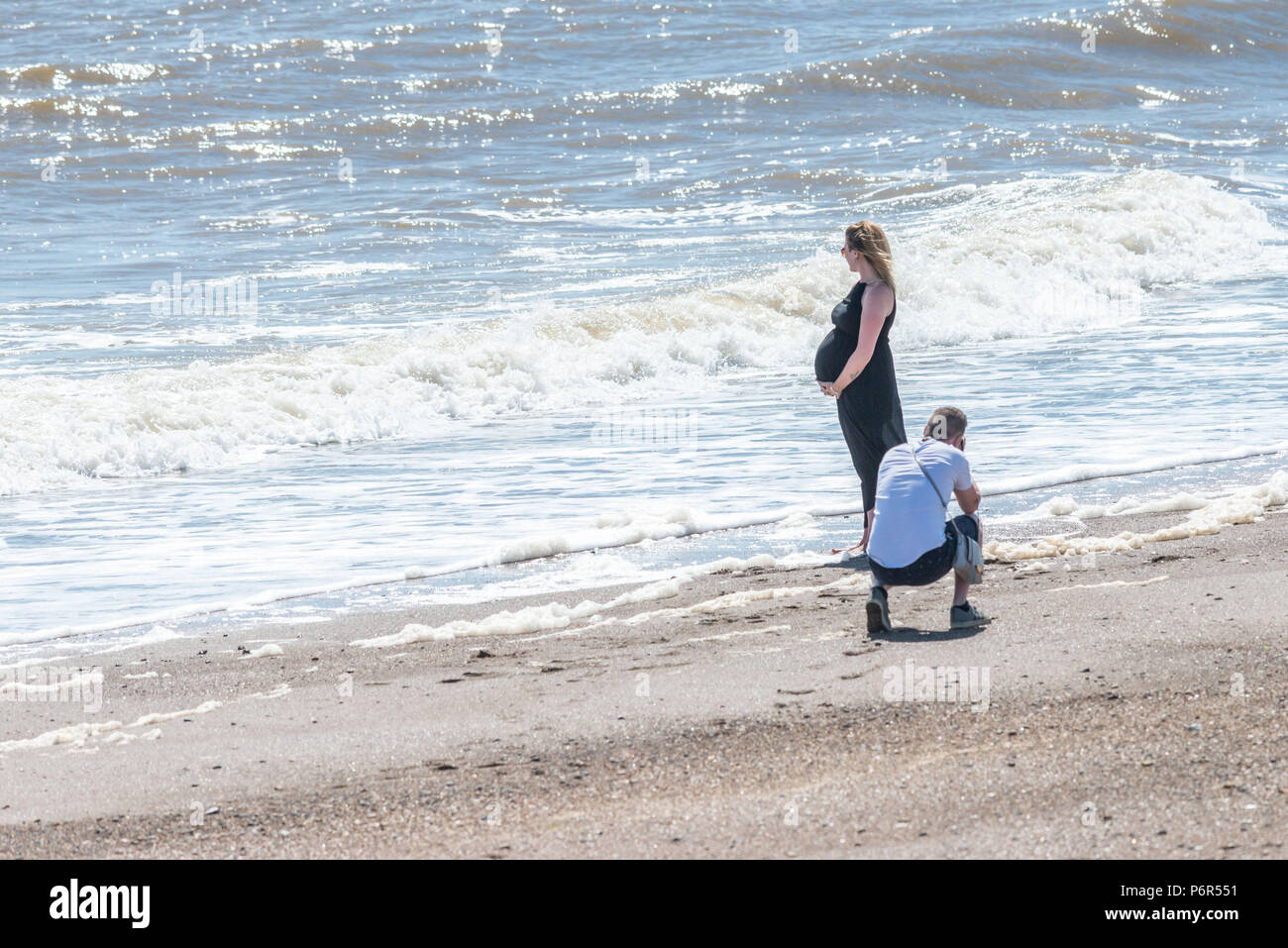 Skegness, UK. 2nd July 2018. A young couple on the beach at Skegness enjoying the current heatwave of hot weather, and temperatures as high as 30 degrees. The man is taking a photo of his pregnant partner while she stands on the beach with the waves at her feet. Credit: Steven Booth/Alamy Live News Stock Photo