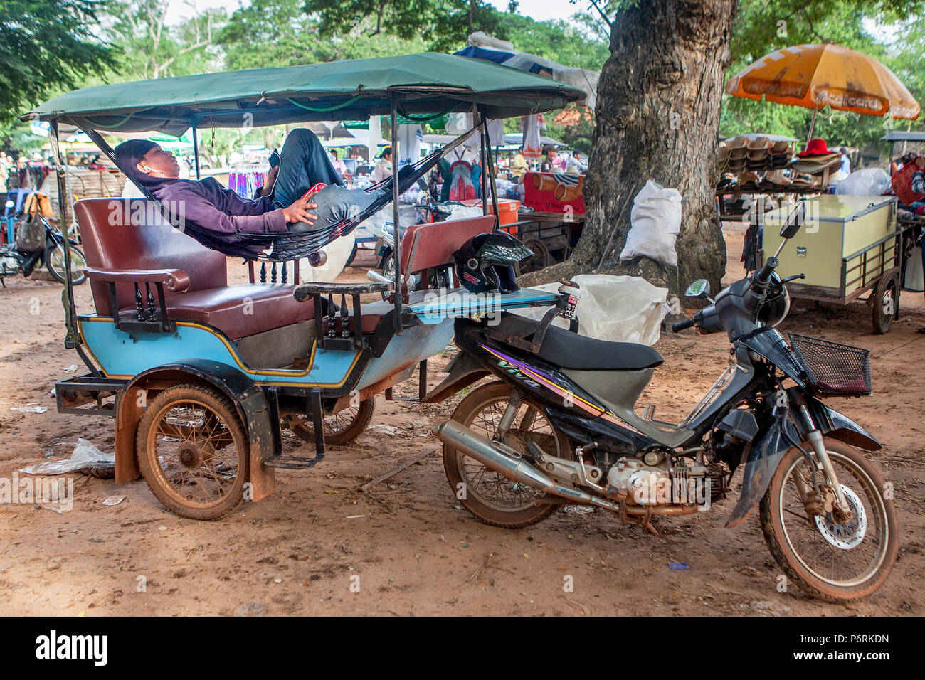A tuk tuk driver relaxes in his hammock listening to music on his smartphone at Angkor Wat, Siem Reap, Cambodia. Stock Photo