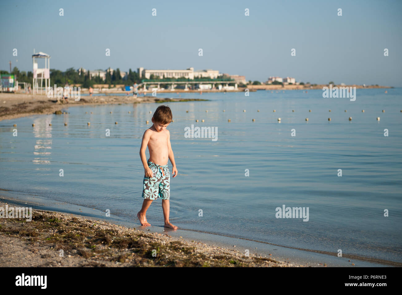 lonely handsome little kid in fashionable shorts walking along sea coast on summer beach Stock Photo