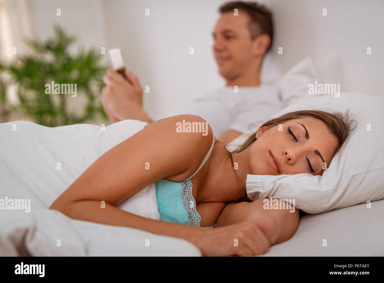 Young woman sleeping while her boyfriend using smartphone in bed. Selective focus. Stock Photo