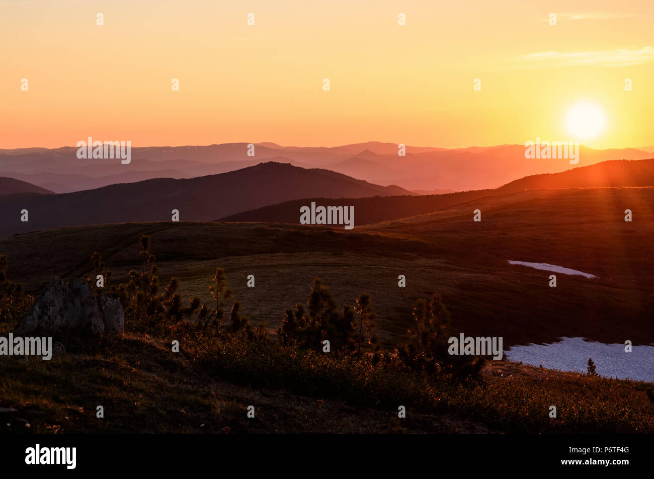 Beautiful sunset with rays over the contours of mountains with stones, small cedars and snow in the foreground Stock Photo