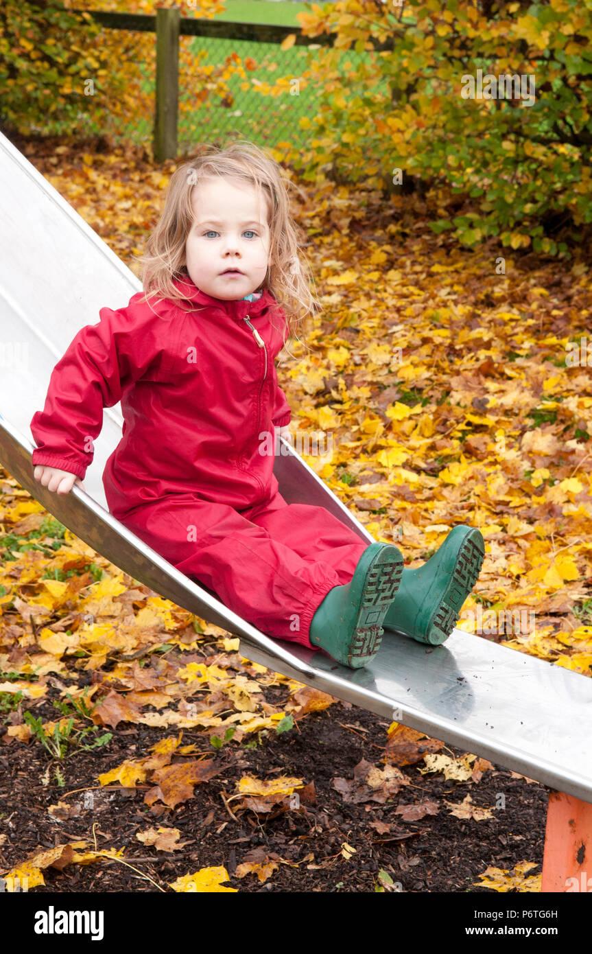 Little girl wearing a red puddle suit and green wellies sitting on a slide Stock Photo