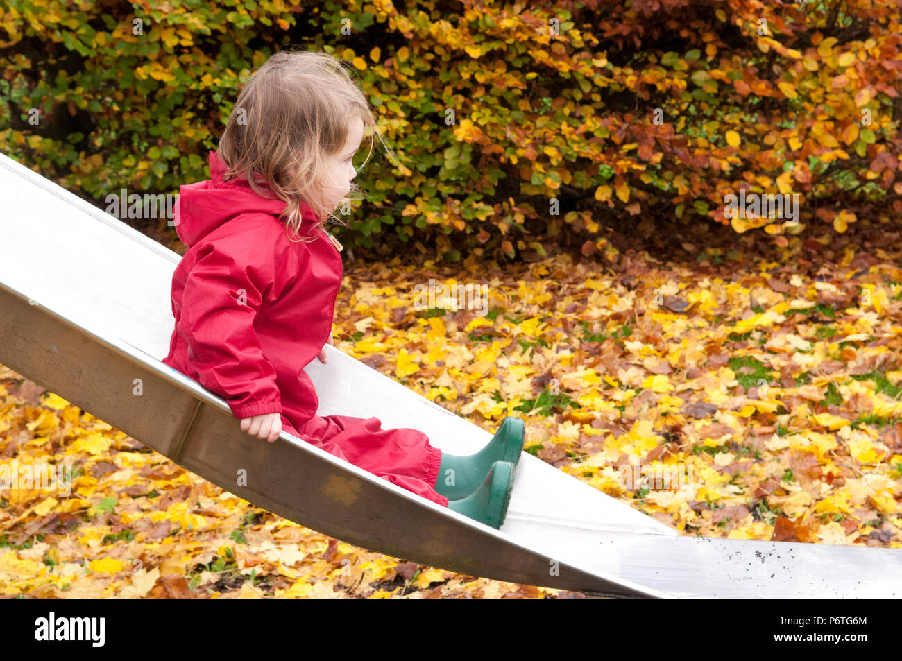 Little girl wearing a red puddle suit and green wellies sitting on a slide Stock Photo