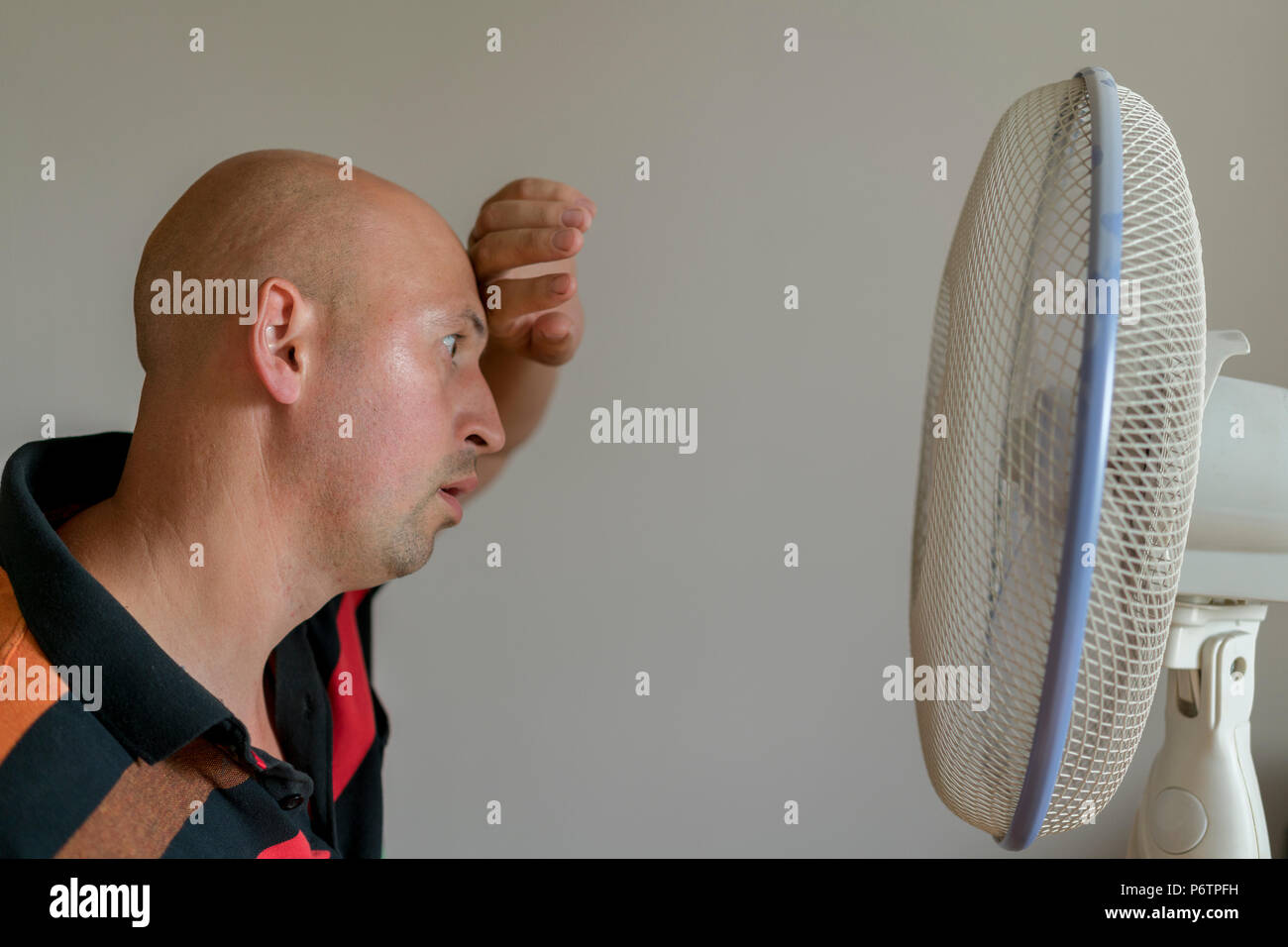 bald man sweating because summer heat haze. Man refreshing in front of air electric fan. Stock Photo