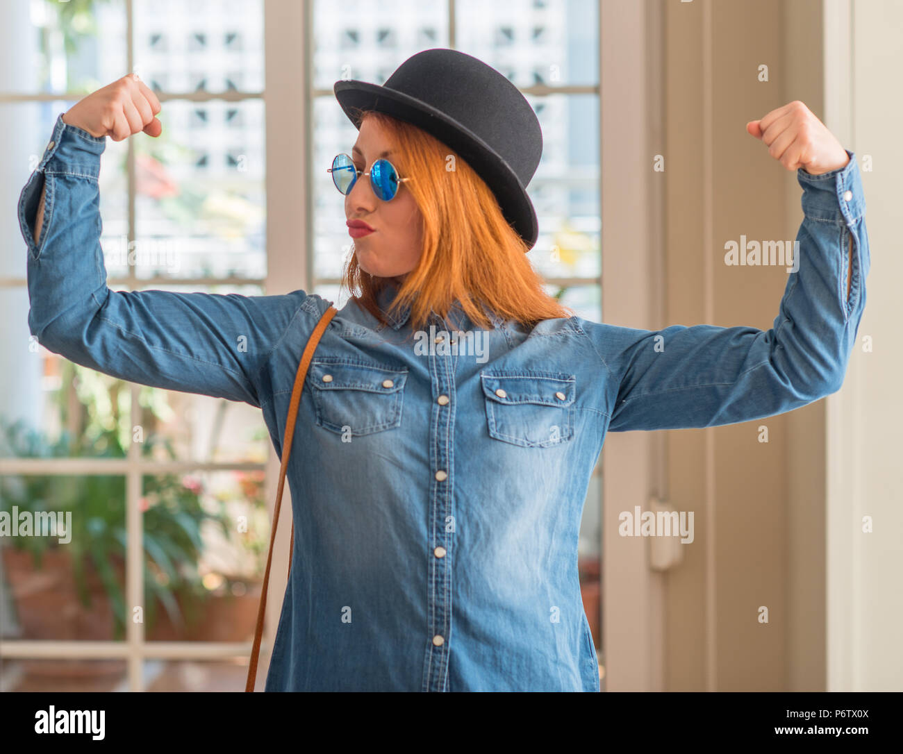 Stylish redhead woman wearing bowler hat and sunglasses showing arms muscles smiling proud. Fitness concept. Stock Photo