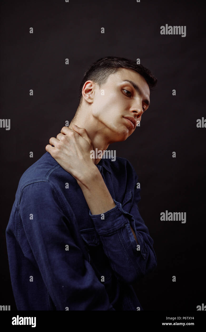 Good-looking upset brown haired young man looking as if he has neck problems or neck pain touching his neck, isolated, over black background Stock Photo