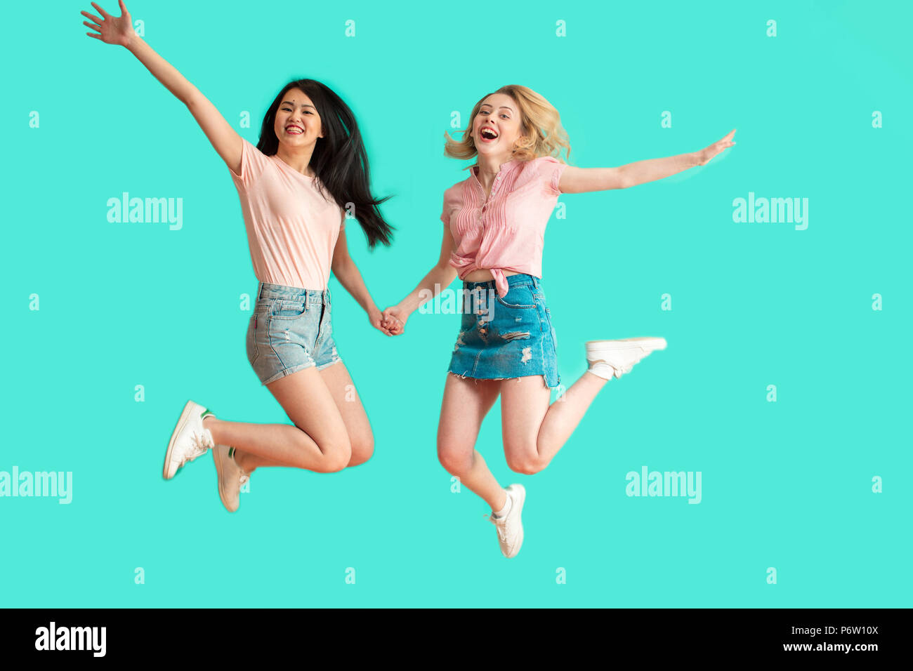 two pretty multiracial brunette and blonde teenage female friends in summer casual wear jumping happy smiling isolated over blue background Stock Photo