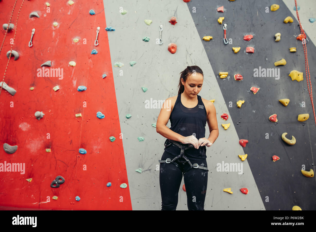 Climbing female instructor ajusting climbing equipment standing over painted in red and grey climbing wall in gym Stock Photo