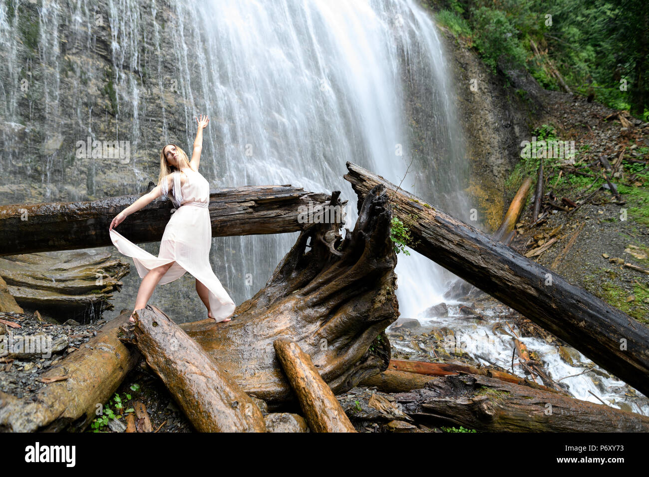 Young woman in pink dress stands barefoot on log, her arms are raised and doing some dreamlike movement and expressing happy feeling at the Bridal Vei Stock Photo