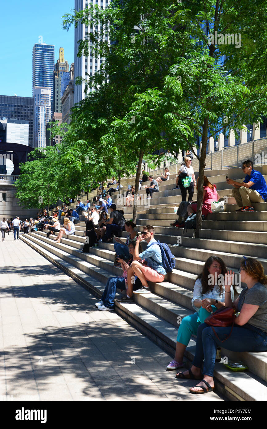 People take a mid day break under the trees on the stadium steps of the Chicago Riverwalk section known as the River Theater. Stock Photo
