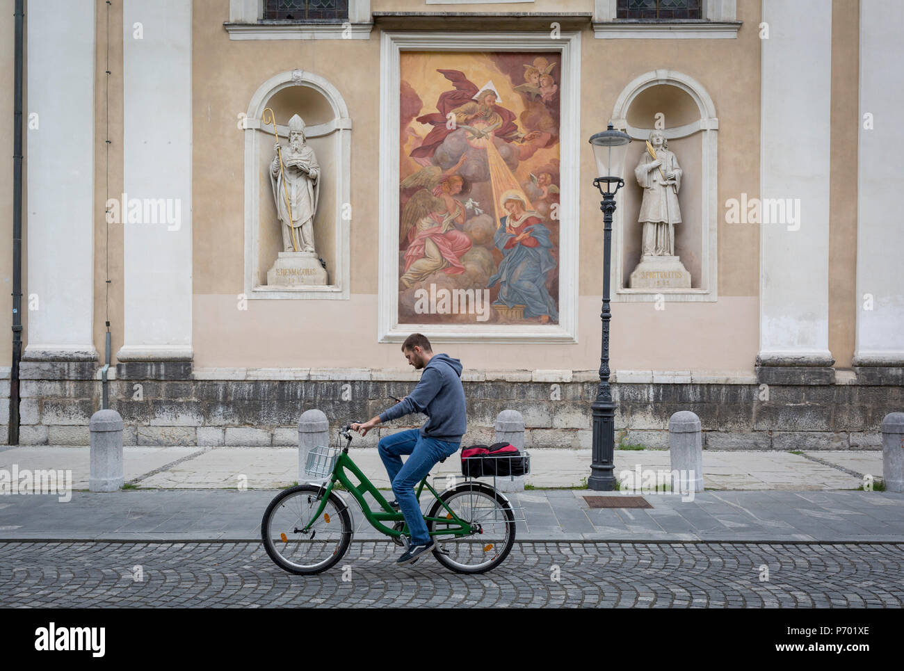 While checking his phone, a cyclist rides past a mural and sculpture outside Cathedral of saint Nicholas in the Slovenian capital, Ljubljana, on 28th June 2018, in Ljubljana, Slovenia. Ljubljana is a small city with flat terrain and a good cycling infrastructure. It was featured at eighth on the 'Copenhagenize' index listing the most bike-friendly cities in the world though bike theft is prevalent. Stock Photo