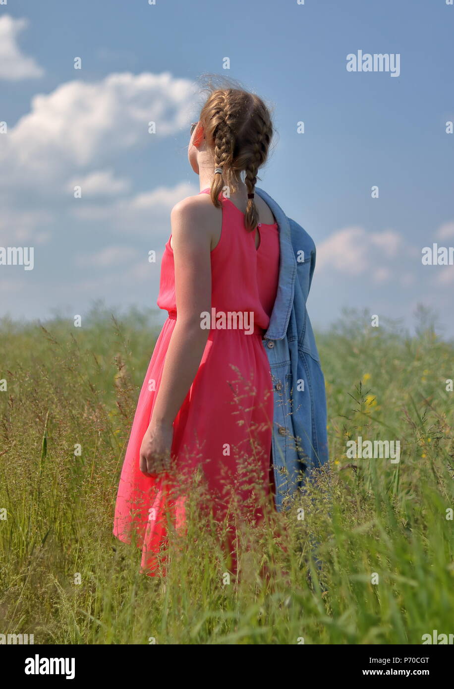 One white skin young blond hair girl stands back on meadow dressed in pink summer sleevless dress, sunny weather, blue sky whith nice white clouds Stock Photo