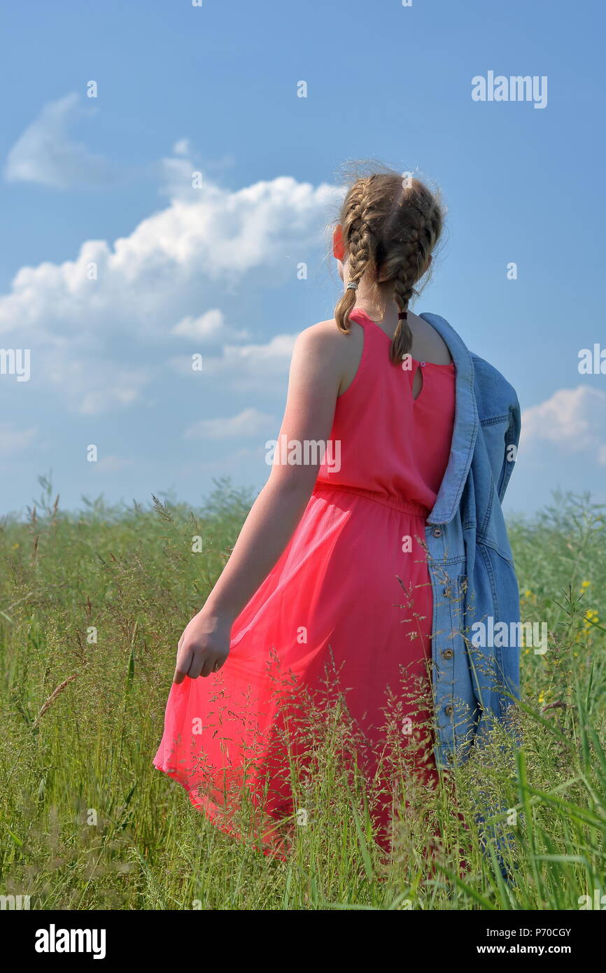 Young blond hair (short braids) teenager girl stand back in meadow, dressed in pink summer sleeveless dress, holds blue jeans jacket on her arm. Stock Photo