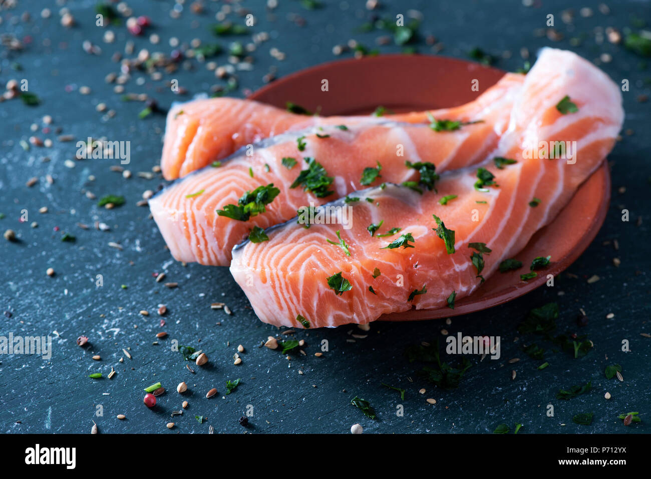 closeup of some slices of raw salmon on a white marble tray, on a dark rustic surface sprinkled with herbs and spices Stock Photo