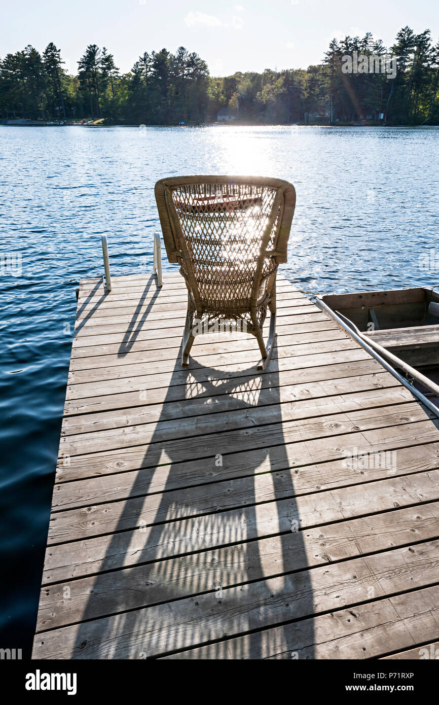 Wicker rocking chair on wooden dock in summer at small lake casting long shadow Stock Photo