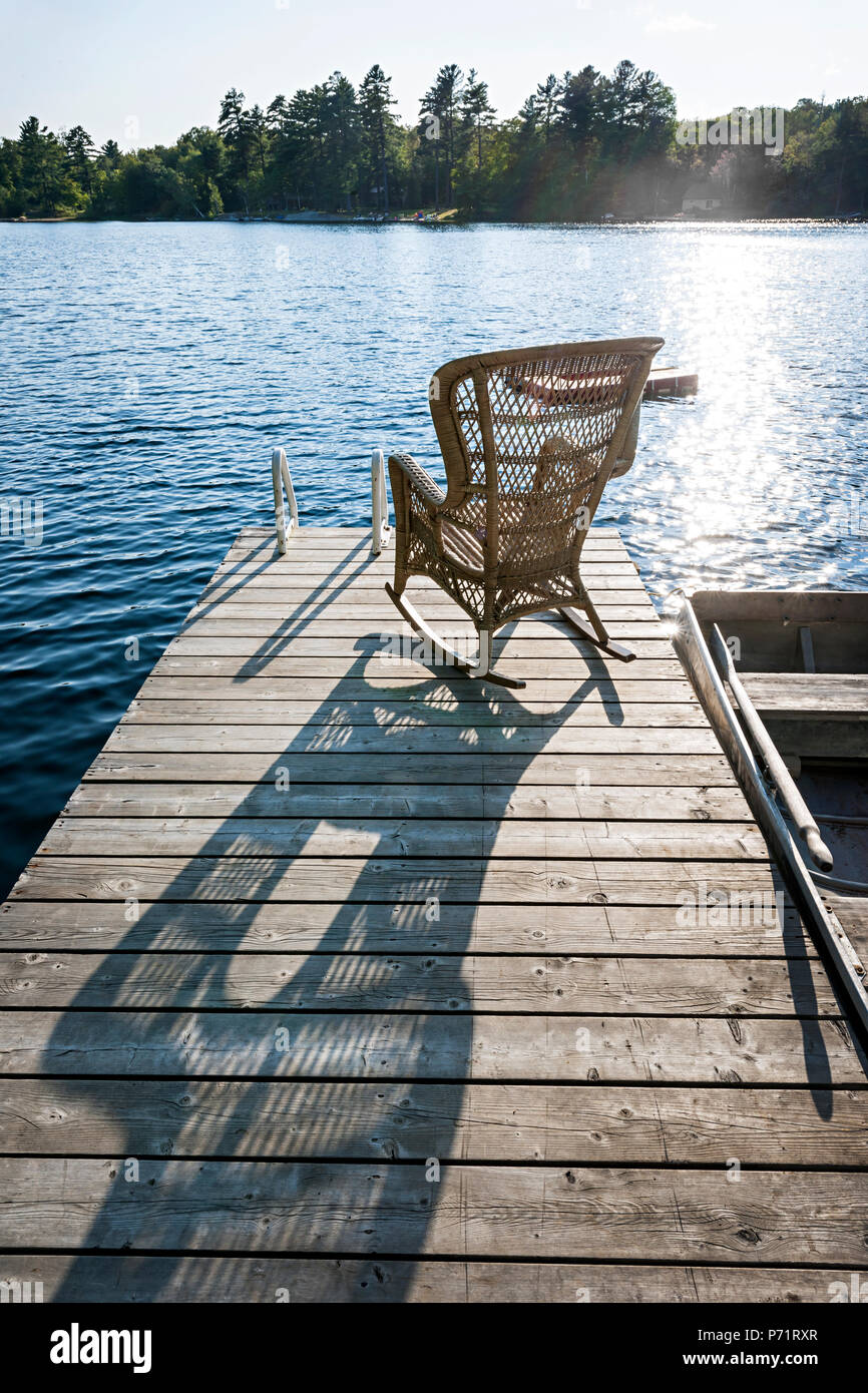 Wicker rocking chair on wooden dock in summer at small lake casting long shadow Stock Photo